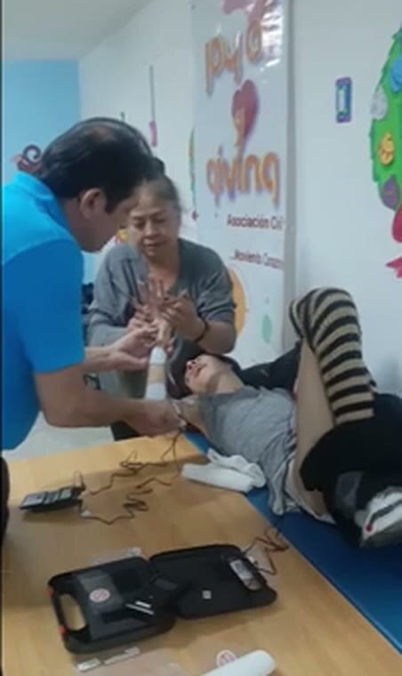 A therapist works with Manuel's hand while a family member supports him. The electro-stimulation unit sits open on the table beside them.