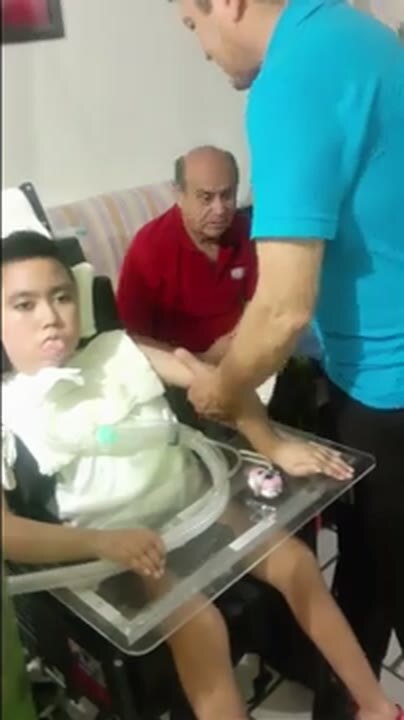 Jaime seated in a supportive chair while a therapist works with his arm, with a family member beside him.